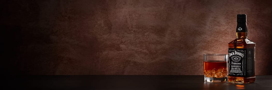 St.Petersburg, Russia - December 2019 - Bottle of Jack Daniel's whiskey and glass with drink and ice on wooden table on brown background with copy space. Extra wide panorama banner background