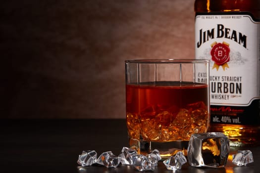St.Petersburg, Russia - December 2019 - Bottle of Jim Beam bourbon whiskey and glass with drink and ice  on wooden table on brown background with copy space. Kentucky straight bourbon whiskey