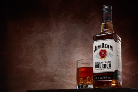 St.Petersburg, Russia - December 2019 - Bottle of Jim Beam bourbon whiskey and glass with drink and ice  on wooden table on brown background with copy space. Kentucky straight bourbon whiskey