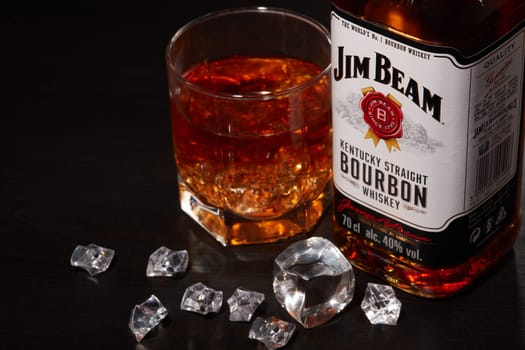 St.Petersburg, Russia - December 2019 - Bottle of Jim Beam bourbon whiskey and glass with drink and ice  on wooden table on brown background. Kentucky straight bourbon whiskey