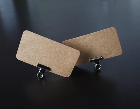 Two blank kraft business cards and metal binder clips on black wooden background. Mockup for branding identity.