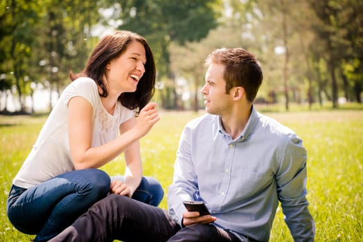 Young woman blowing dandelion to face of her boyfriend holding mobile phone