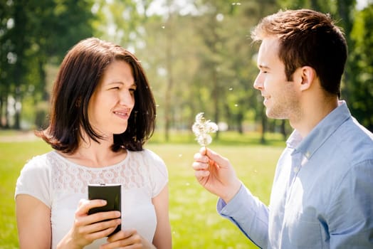Young man blowing dandelion to face of angry woman holding mobile phone