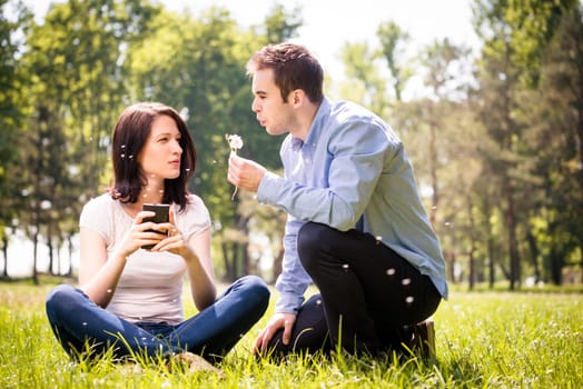 Young man blowing dandelion to face of angry woman holding mobile phone