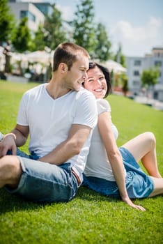 Young couple sitting on grass, leaning with backs and smiling over shoulder
