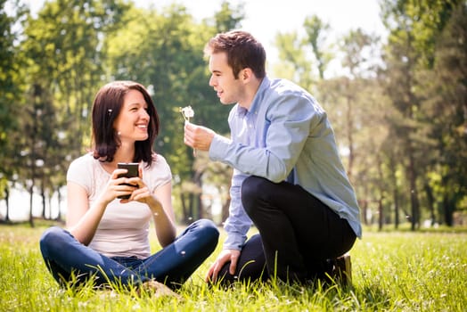 Young man blowing dandelion to face of his girlfriend holding mobile phone