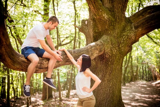Young man sitting on big tree invites his girlfriend standing bellow