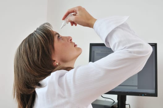 Detail of young business woman applying eye drops on workplace - computer monitor in background