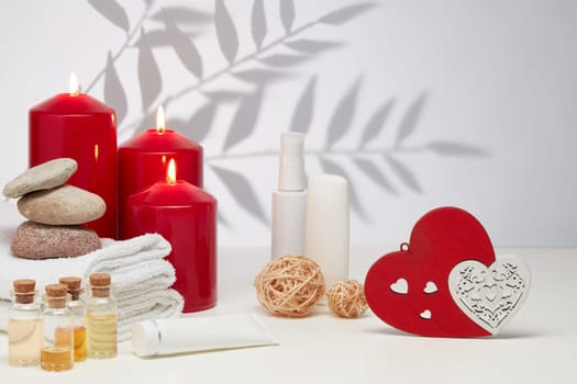Spa still life with creams, essential oils, burning red candles, hearts on light background with shadow from plant. Healthy lifestyle, body care, Spa treatment. Valentine's Day. Honeymoon. Wedding