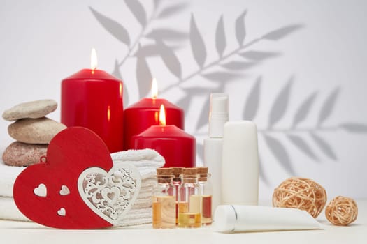 Spa still life with creams, essential oils, burning red candles, hearts on light background with shadow from plant. Healthy lifestyle, body care, Spa treatment. Valentine's Day. Honeymoon. Wedding