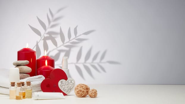 Spa still life with creams, essential oils, burning red candles, hearts on light background with shadow from plant. Healthy lifestyle, body care, Spa treatment. Valentine's Day. Honeymoon. Wedding