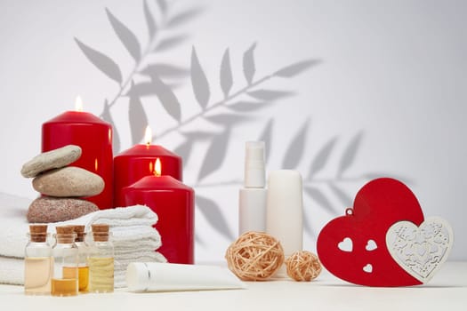Spa still life with creams, essential oils, burning red candles, hearts on light background with shadow from plant. Healthy lifestyle, body care, Spa treatment. Valentine's Day. Honeymoon. Wedding