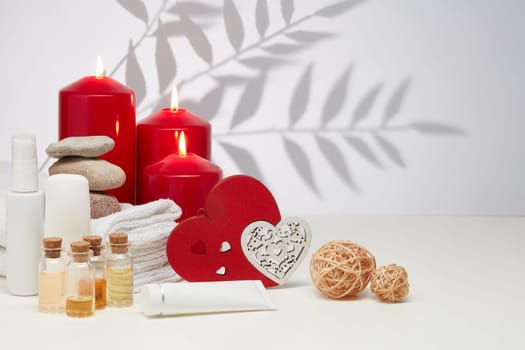 Spa still life with creams, essential oils, burning red candles, hearts on light background with shadow from plant. Healthy lifestyle, body care, Spa treatment. Valentine's Day. Honeymoon. Wedding