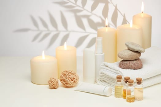 Spa still life with creams, essential oils, burning candles, stones and towels on light background with shadow from plant. Healthy lifestyle, body care, Spa treatment.