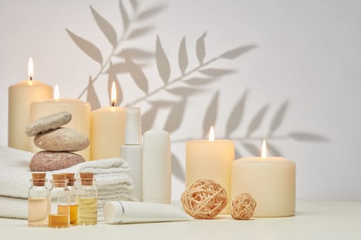Spa still life with creams, essential oils, burning candles, stones and towels on light background with shadow from plant. Healthy lifestyle, body care, Spa treatment.