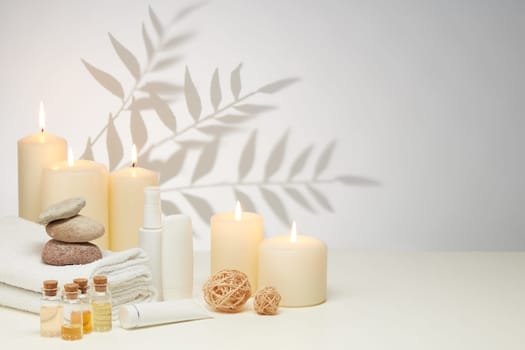 Spa still life with creams, essential oils, burning candles, stones and towels on light background with shadow from plant. Healthy lifestyle, body care, Spa treatment.