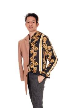 A tall skinny young man in a gold printed shirt and his hand in his.pocket, with beige coat over his shoulder, isolated for white background.