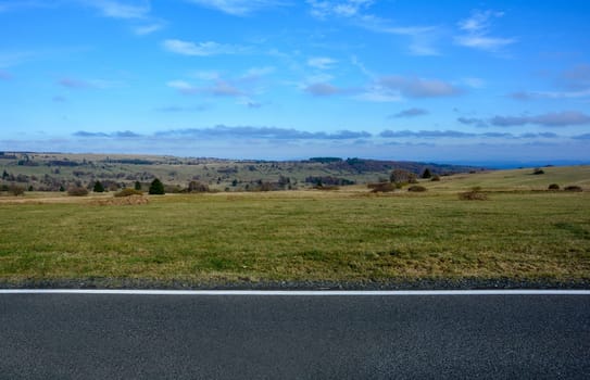 Landscape with part of a street in the lower part of the picture, with a large meadow, trees and blue sky in the Rhön, Bavaria, Germany