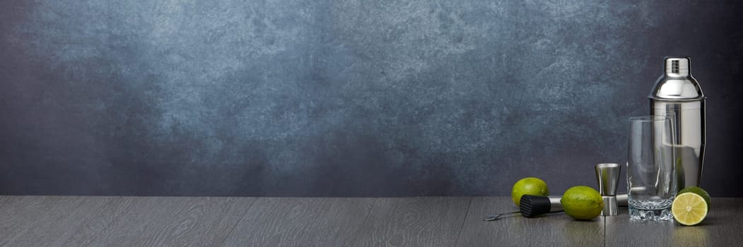 Set of bar tools: cocktail shaker, muddler, jigger, bar spoon, empty highball glass and  limes on dark gray background with copy space. Bartender's tool.