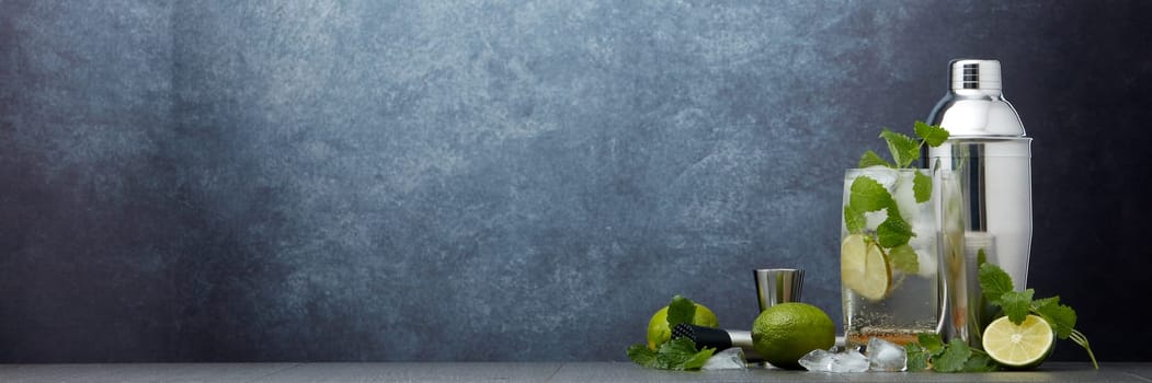 Set of bar tools: cocktail shaker, muddler, jigger, cocktail with ice in highball glass,  lime and mint on dark gray background with copy space. Bartender's tool. Mojito cocktail making.