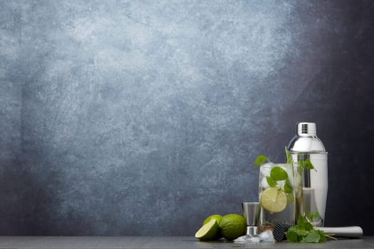 Set of bar tools: cocktail shaker, muddler, jigger, cocktail with ice in highball glass,  lime and mint on dark gray background with copy space. Bartender's tool. Mojito cocktail making.