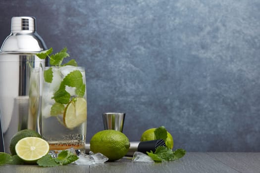 Set of bar tools: cocktail shaker, muddler, jigger, cocktail with ice in highball glass,  lime and mint on dark gray background with copy space. Bartender's tool. Mojito cocktail making.
