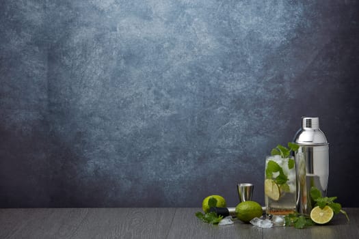 Set of bar tools: cocktail shaker, muddler, jigger, cocktail with ice in highball glass,  lime and mint on dark gray background with copy space. Bartender's tool. Mojito cocktail making.
