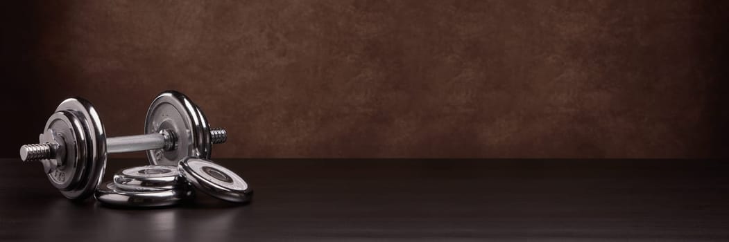Steel dumbbell and weights on dark wooden surface on brown background with copy space. Sports bodybuilding equipment. Fitness, sport, healthy lifestyle concept. Extra wide panorama banner background