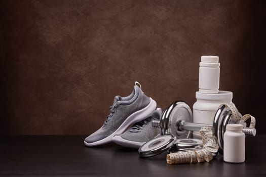 White plastic containers with sports nutrition (supplements), measuring tape, sneakers, dumbbells on dark background with copy space. Fitness, bodybuilding, slimming, sport, healthy lifestyle concept.