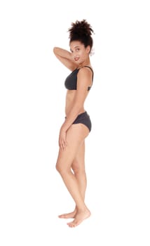 A beautiful slim young multi-racial woman standing in profile in a black.bikini in the studio looking into camera, bare foot with her curly black .hair in a bun on her head, isolated for white background.