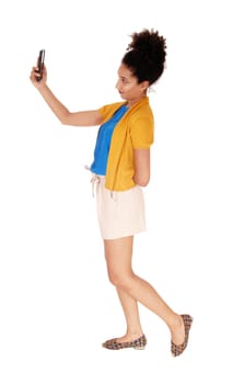 Beautiful young multi-racial woman standing in shorts and a yellow.sweater in profile looking at cellphone isolated for white background.