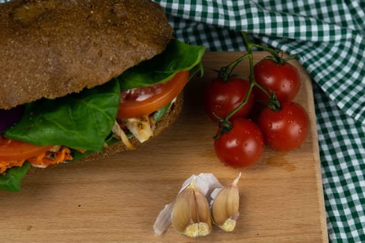 A dark bread sandwich ut in half with chicken, tomatoes, spinach, cheese and carrots stands on a wooden board that stands on a checkered white green towel. Top views