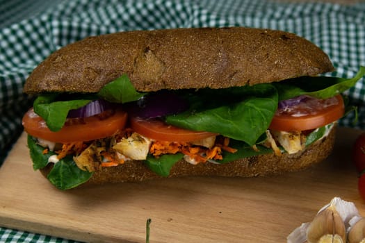A dark bread sandwich ut in half with chicken, tomatoes, spinach, cheese and carrots stands on a wooden board that stands on a checkered white green towel. Top views