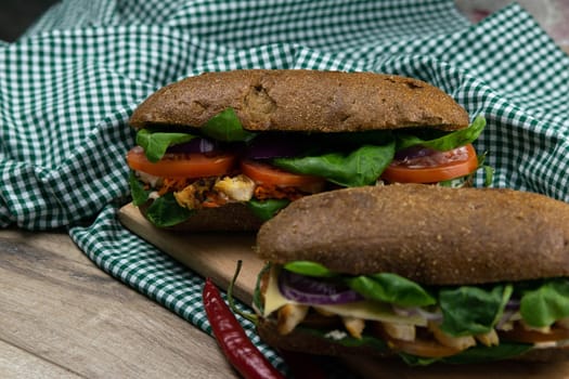 A dark bread sandwich ut in half with chicken, tomatoes, spinach, cheese and carrots stands on a wooden board that stands on a checkered white green towel. Top views
