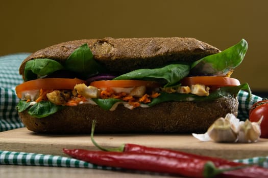 A dark bread sandwich ut in half with chicken, tomatoes, spinach, cheese and carrots stands on a wooden board that stands on a checkered white green towel. Top views