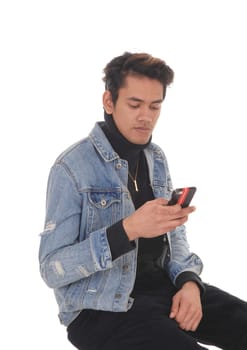 A young handsome man sitting in a jeans jacket and black pants.on a chair, looking at his cell phone, isolated for white background.