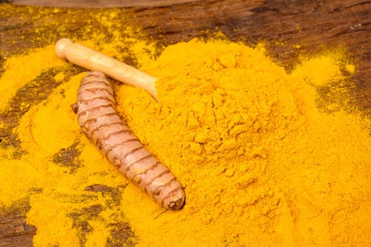 Dried curcuma powder with a wooden spoon and a whole fresh turmeric root in fornt of a wooden background.