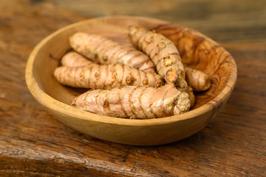 Fresh whole turmeric roots in a wooden bowl in front of a wooden rustic background.