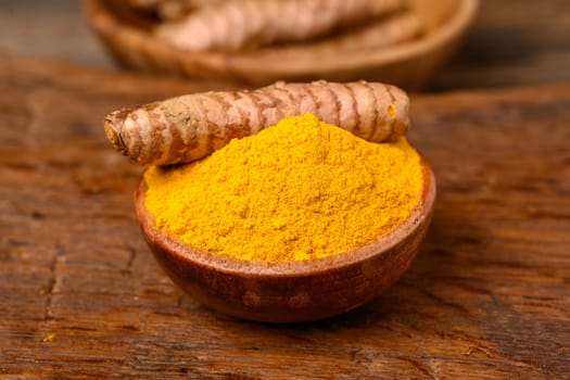 A wooden bowl with curcuma powder and one whole turmeric root lies on top in front of a wooden background.