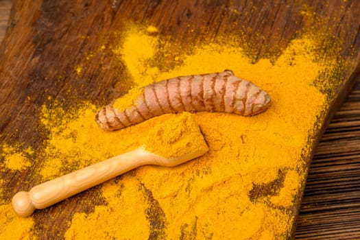 Dried curcuma powder with a wooden spoon and a whole fresh turmeric root in front of a wooden background.