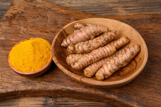 A wooden bowl with curcuma powder and a bowl with whole turmeric roots on a wooden cutting board.