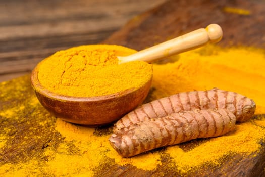 Dried curcuma powder in a wooden bowl with a wooden spoon and whole fresh turmeric roots in front of a wooden background.