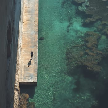 A lone person walks on a sunlit stone pier beside transparent turquoise water, casting a long shadow in a peaceful and minimalist coastal scene.