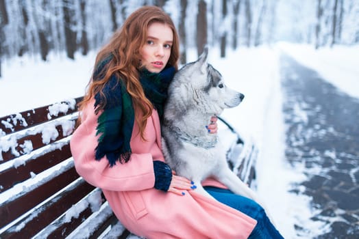 Beautiful woman sitting on the bench with siberian husky, snowy forest on background. Cute girl hugs with charming dog. Real friendship with domestic animal