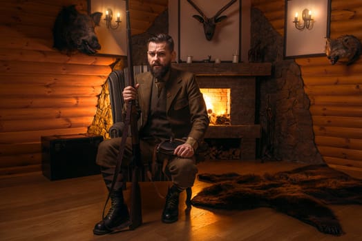 Vintage hunter man in traditional hunting clothing sitting in a chair with retro rifle against burning fireplace. Stuffed wild animals, bear skin and other trophies on background