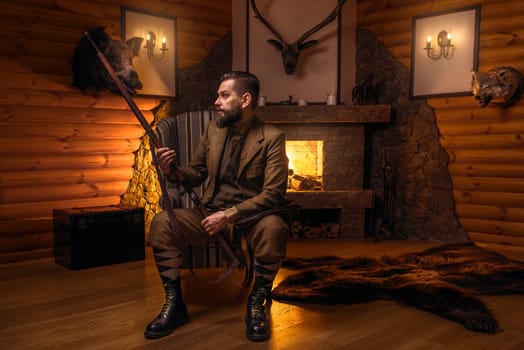 Vintage hunter man in traditional hunting clothing sitting in a chair with retro rifle against burning fireplace. Stuffed wild animals, bear skin and other trophies on background
