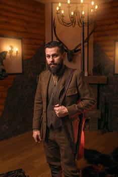 Hunter man with old gun in vintage traditional hunting clothing standing against antique chest. Fireplace, stuffed wild animals, bear skin and other trophies on background