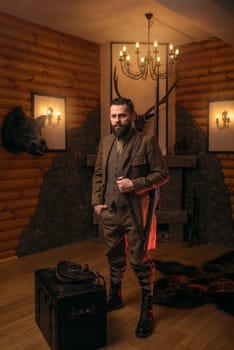 Hunter man with old gun in vintage traditional hunting clothing standing against antique chest. Fireplace, stuffed wild animals, bear skin and other trophies on background