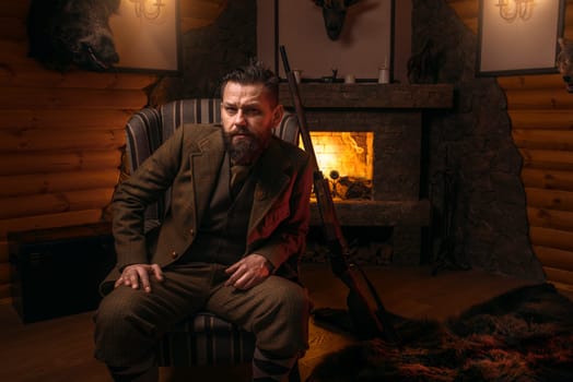 Solid male hunter in traditional hunting clothes sitting in a chair against burning fireplace. Retro style, vintage rifle. Stuffed wild animals, bear skin on background