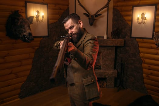 Hunter man aims of the antique hunting rifle. Fireplace, stuffed wild animals, bear skin and other trophies on background
